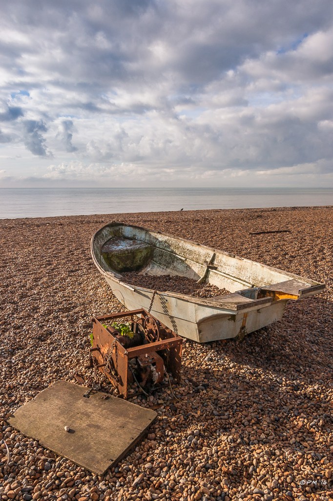 Old rowing boat chained to rusty winch and plywood board sitting on Brighton Beach, UK 2014 P.Maton eyeteeth.net