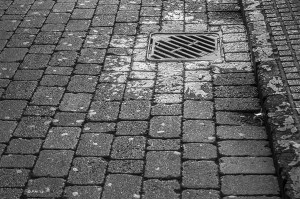 Wet drain on stone path lit by morning sun. Monochrome abstract. Brighton East Sussex UK. P.Maton 2014 eyeteeth.net