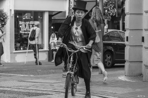 Oriental lady in top hat and black coat and skirt riding small bicycle. Monochrome landscape. Gardener Street, Brighton, UK © P.Maton 2014 eyeteeth.net