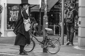Oriental lady in top hat and black coat and skirt standing next to small bicycle. Monochrome landscape. Gardener Street, Brighton, UK © P.Maton 2014 eyeteeth.net