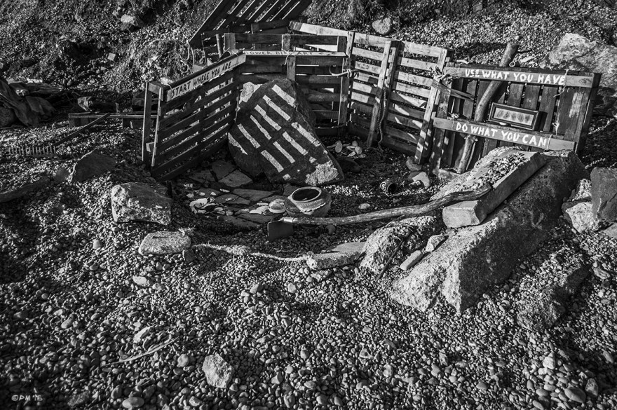 Beachcomber's den mad from pallets on shingle beach with makeshift tool and collected items and signs saying - Start Where You Are, Use What You Have, Do What You Can. Shoreham Beach Sussex UK. Monochrome Landscape. © P. Maton 2015 eyeteeth.net