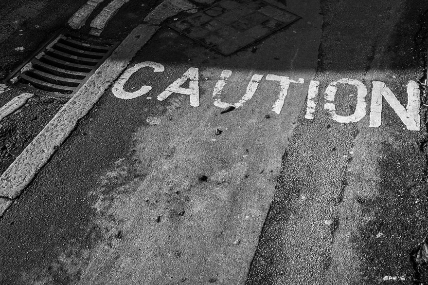 Caution notice written on pavement / sidewalk next to raod with drain grille and shadow. Viaduct Road, Brighton UK. Monochrome Landscape. © P. Maton 2015 eyeteeth.net