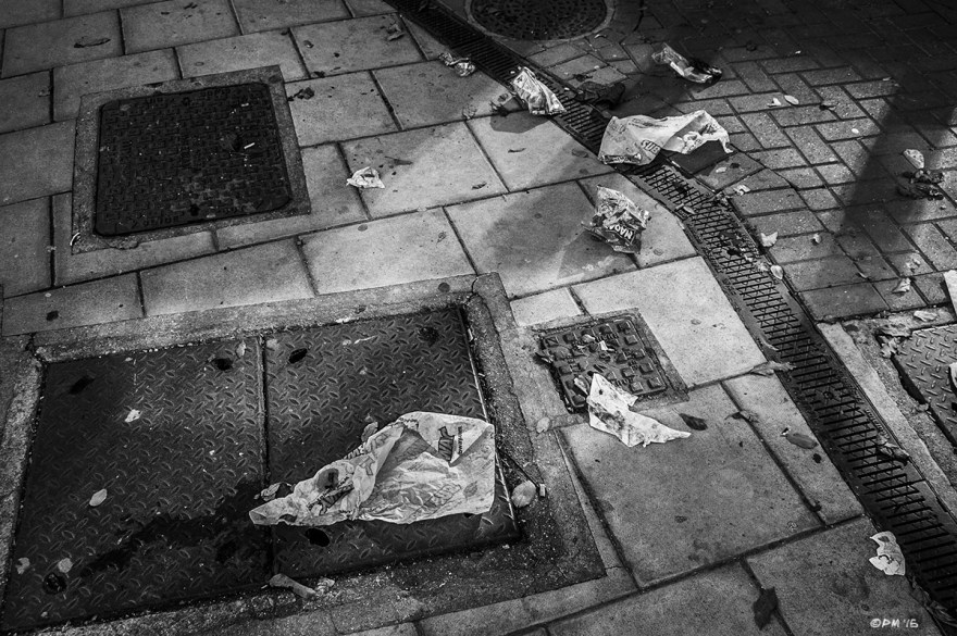 Drain covers and service wells in pavement with litter / trash and reflected light. Edward Street, Brighton UK. Monochrome Landscape. © P. Maton 2015 eyeteeth.net