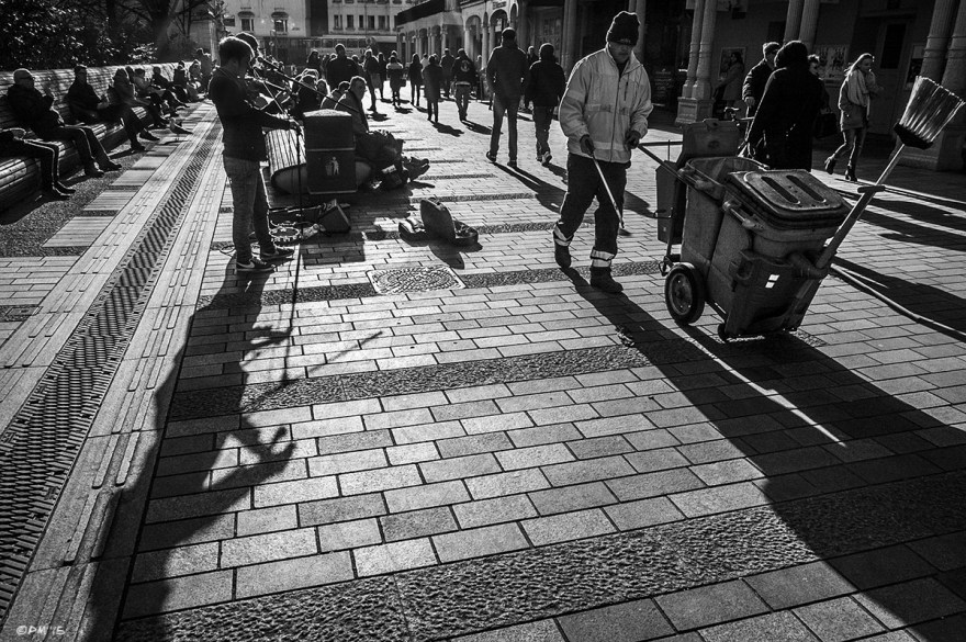 Buskers and street sweeper among people walking down paved street silhouetted by winter sun. New Road, Brighton UK. Monochrome Landscape. © P. Maton 2015 eyeteeth.net