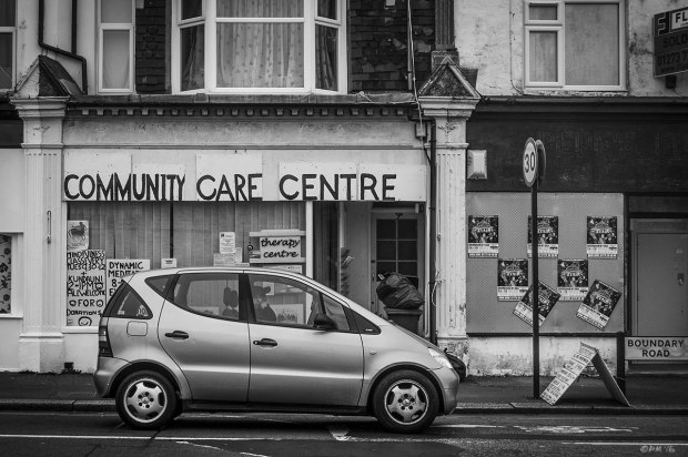 Shop front with hand painted sign saying Community Care Centre and shuttered empty shop with care in foreground. Boundary Road, Portslade On Sea, Sussex UK.  Monochrome Landscape. © P. Maton 2015 eyeteeth.net