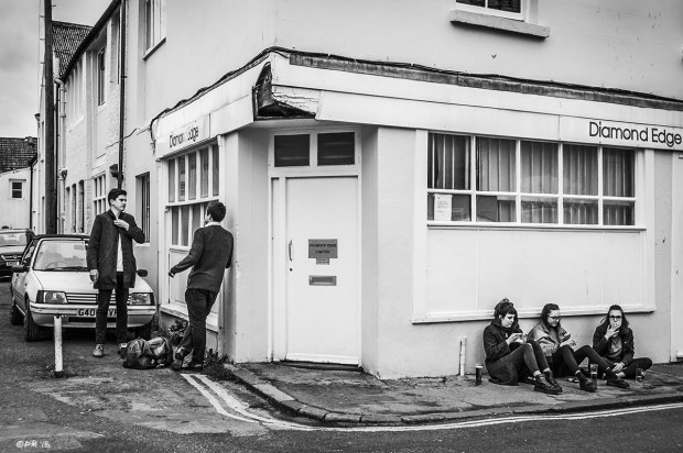Two young me standing on street corner by three young women sitting on pavement smoking. Gloucester Road Brighton UK.  Monochrome Landscape. © P. Maton 2015 eyeteeth.net