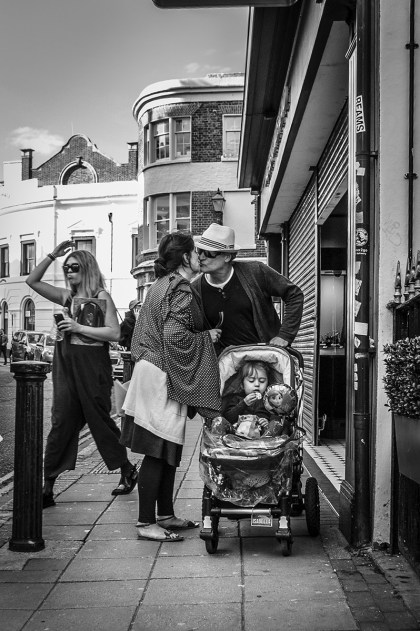 Couple with pram kissing in street while child looks at a potato chip / crisp with woman raising her arm in background. Ship Street Brighton UK. Ship Street Brighton UK. Street Photography. Monochrome Portrait. © P. Maton 2015 eyeteeth.net