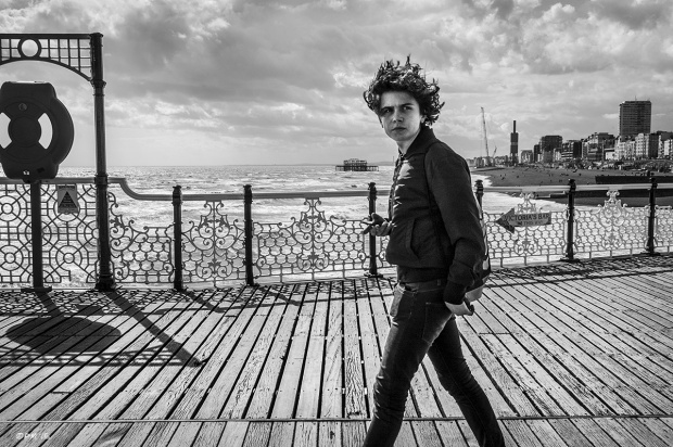 Young man looking passed viewer, walling along brighton pier with hair blowing in the wind . View of sea front and West Pier in background. Palace Pier Brighton UK. Monochrome Landscape. © P. Maton 2015 eyeteeth.net