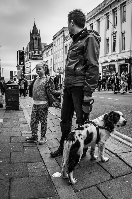 Girl putting on coat while father looks down street towards sea holding spaniel on a lead. West Street Brighton Sussex UK. Black and white urban street photograph. © P. Maton 2016 eyeteeth.net.