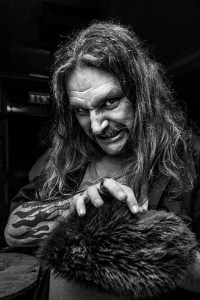 moustached man with long hair, tattoos and claw like hand reaching over a fake fur hat while grimacing in a sinister fashion at the camera. Black and white portrait. © P. Maton 2016 eyeteeth.net