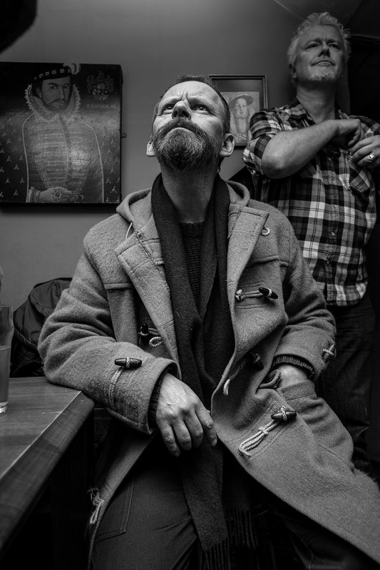 Man with beard in duffel coat looking upwards into light, Shakespeares Head Pub Brighton UK. Black and white nightlife documentary portrait photography. © P. Maton 2017 eyeteeth.net