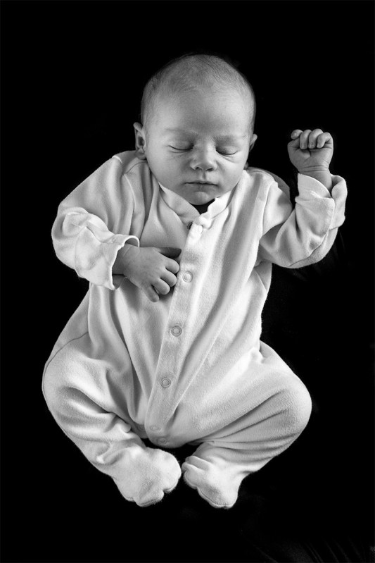 Sleeping baby in romper suit against black background with fist raised. Black and white portrait photography. Fuji film XT-2 23mm f2 R WR. © P. Maton 2017 eyeteeth.net