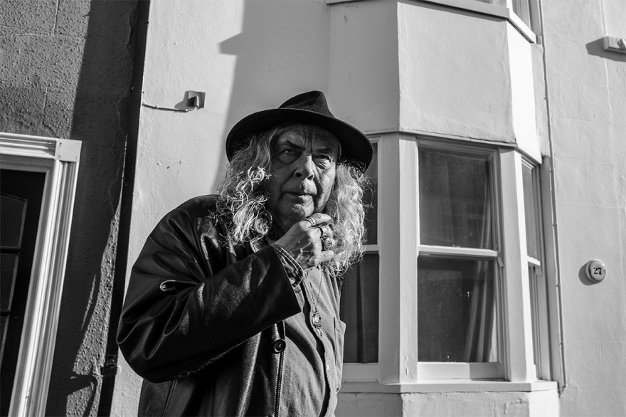 Smoking man with long curly hair and fedora hat in front of houses on Cheltenham Place Brighton UK.  Urban street photography Black and white landscape. © P. Maton 2017 eyeteeth.net
