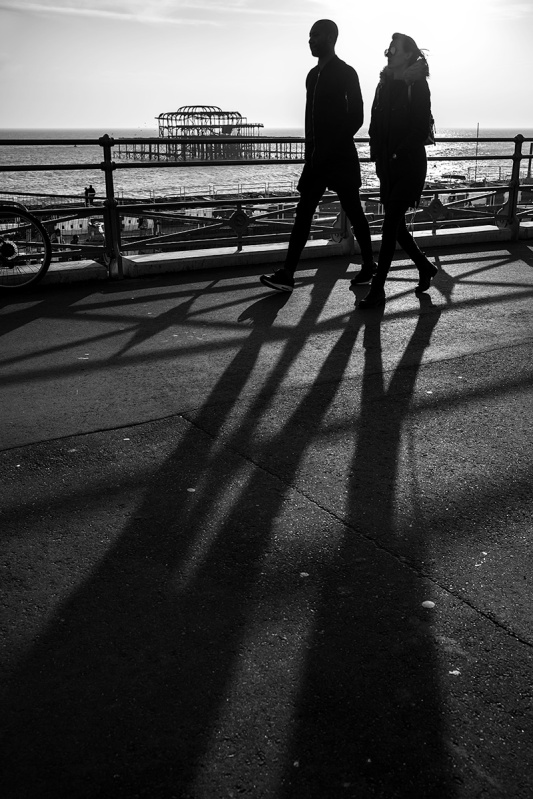Silhouettes couple casting long shadows walking on seafront  Kings Road Brighton with West Pier ruins in background. Black and white urban street photography. © P. Maton 2017 eyeteeth.net