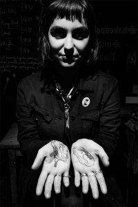 Young woman with black hair dressed in black with palms of hands turned up showing tattoos, black and white high contrast portrait alternative fashion