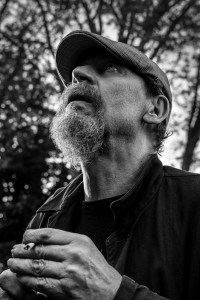 Bearded man wearing flat cap sitting outside holding a cigarette and gazing towards the sky, Shakespeare's Head Pub Brighton UK. Black and white natural light documentary portrait. © P. Maton 2017 eyeteeth.net