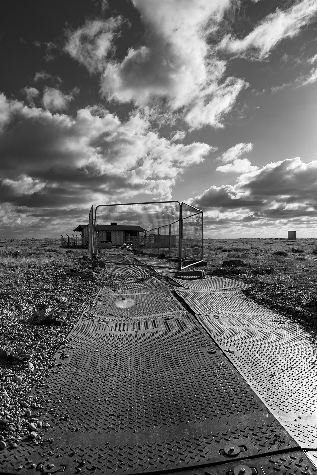 New building construction site, rubber path over shingle with site fencing and dramatic clouds in sky, Dungeness Kent UK, black and white monochrome landscape portrait © P. Maton 2018 eyeteeth.net