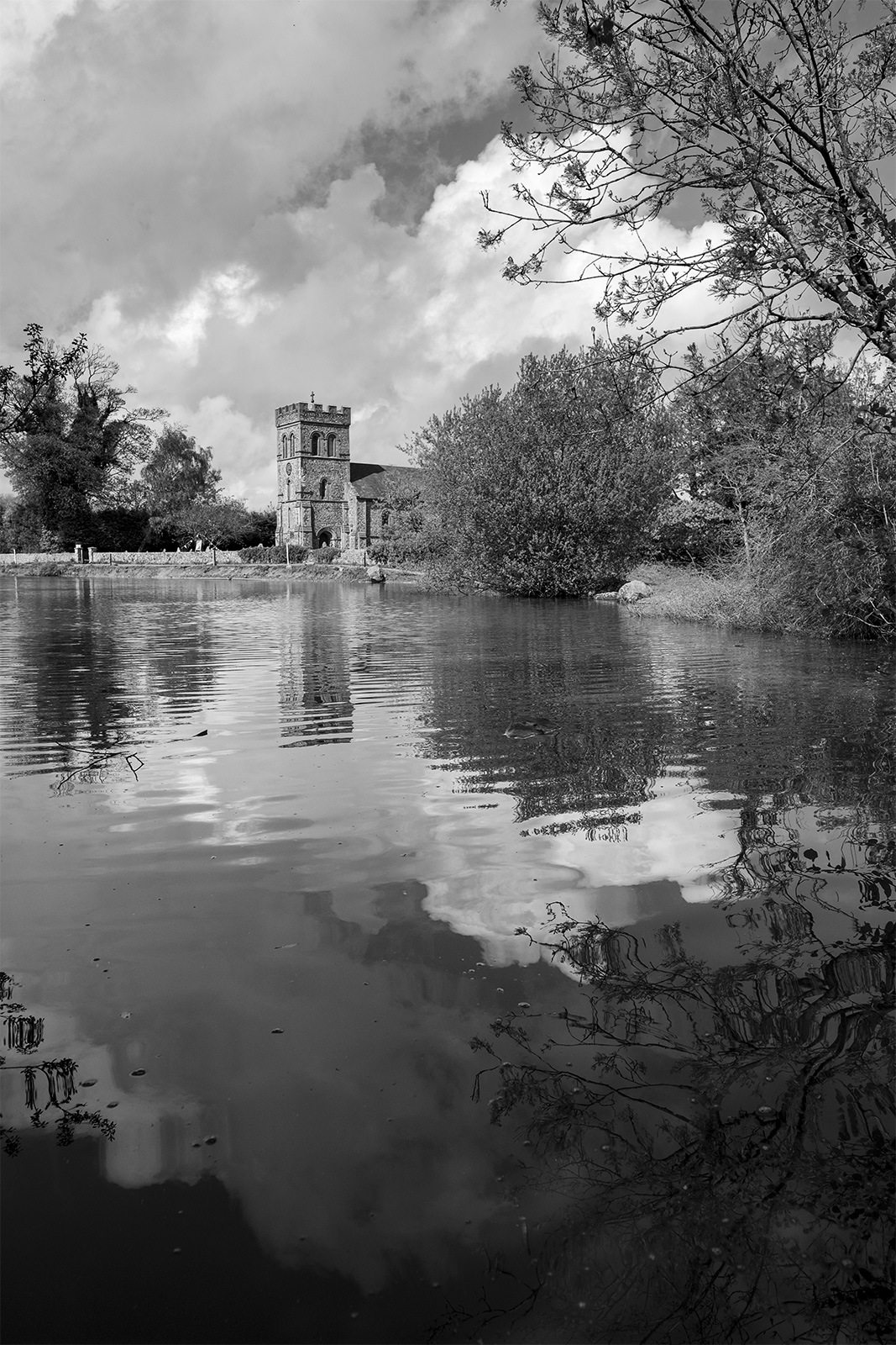 View across pond to Rain Laurence church in Stanmer village East Sussex with clouds in sky UK black and white rural landscape portrait ©P. Maton 2019 eyeteeth.net