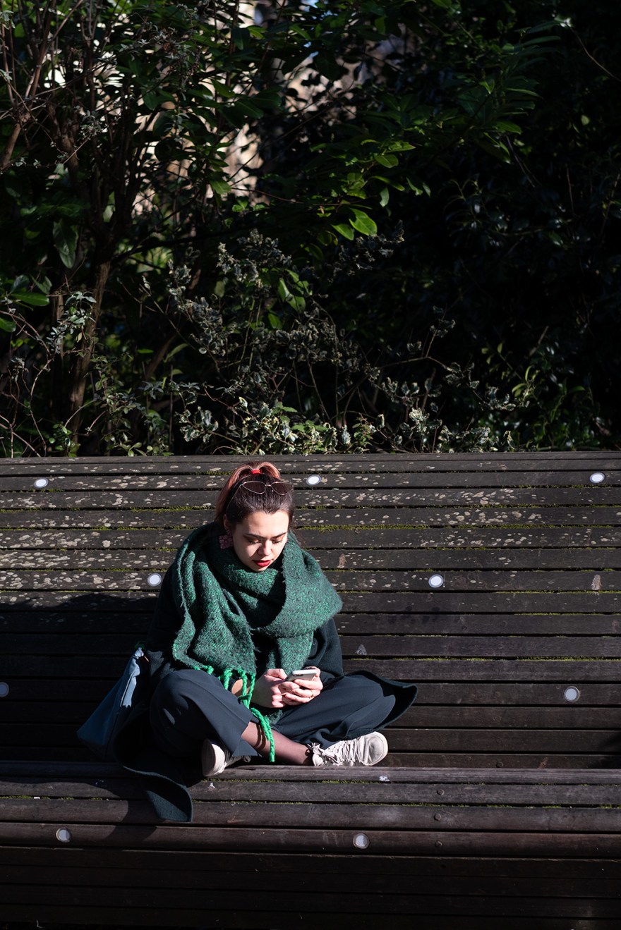 Fashionable stylish young woman sitting with crossed legs on bench studying mobile phone, New Road Brighton UK colour documentary street photography portrait ©P. Maton 2019 eyeteeth.net